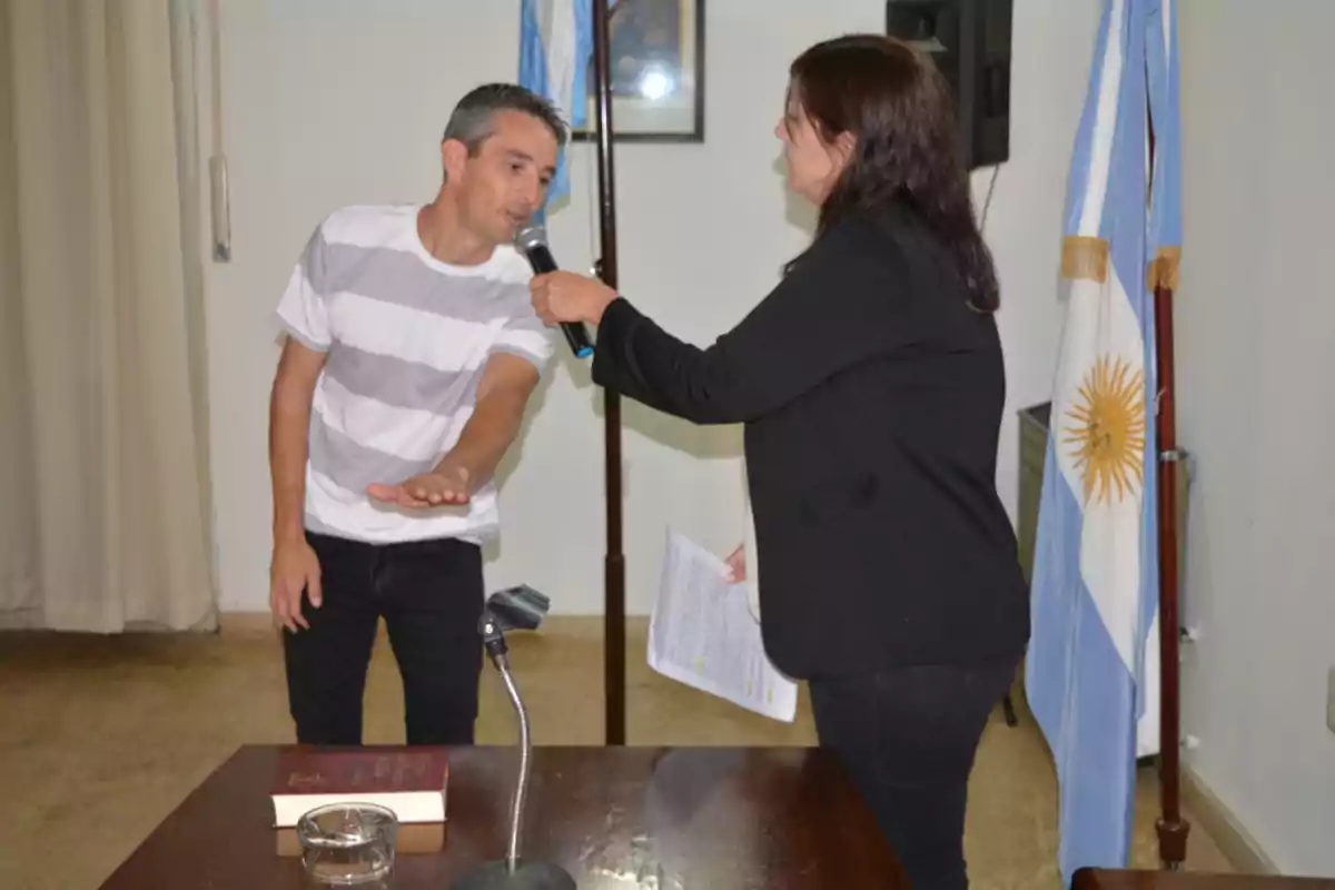 Un hombre con camiseta a rayas está prestando juramento frente a una mujer que sostiene un micrófono, en una sala con banderas argentinas. Un hombre con camiseta a rayas está prestando juramento frente a una mujer que sostiene un micrófono, en una sala con banderas argentinas.