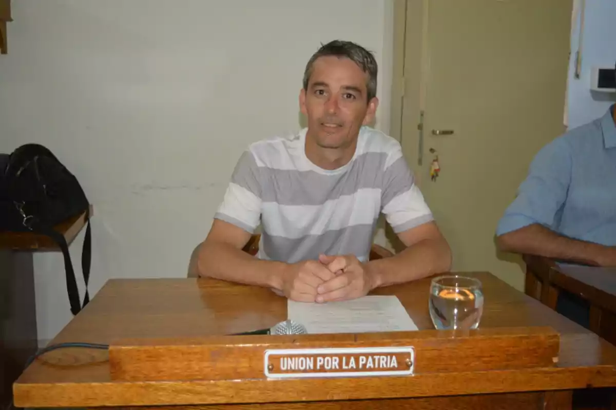 Hombre sentado en un escritorio de madera con un cartel que dice UNION POR LA PATRIA frente a él, acompañado de un vaso de agua, un micrófono y papeles