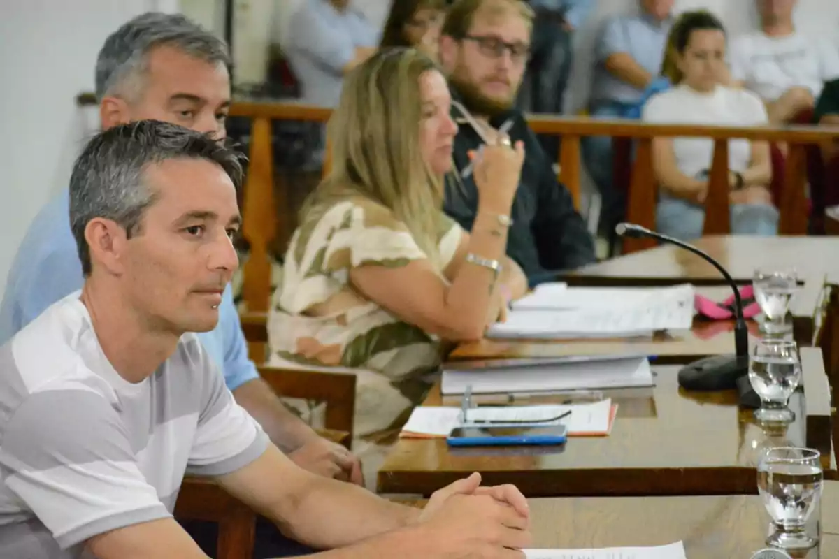 Personas sentadas en una mesa de madera con documentos, micrófonos y vasos de agua, participando en una reunión o sesión formal, con otras personas al fondo observando. Personas sentadas en una mesa de madera con documentos, micrófonos y vasos de agua, participando en una reunión o sesión formal, con otras personas al fondo observando.