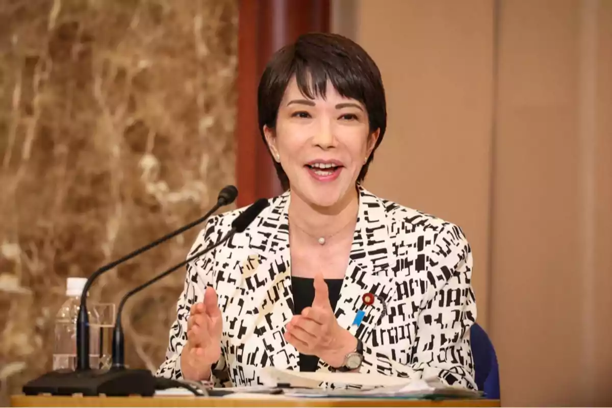 Short-haired woman smiling while speaking in front of two microphones at a table, wearing a black and white blazer, with a marbled wall background. Short-haired woman smiling while speaking in front of two microphones at a table, wearing a black and white blazer, with a marbled wall background.