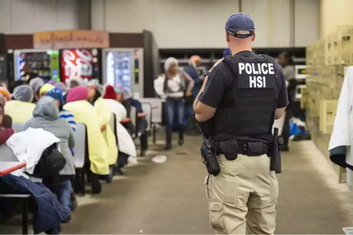 Police officer wearing a vest labeled HSI supervises a group of people seated in a room with vending machines in the background.
