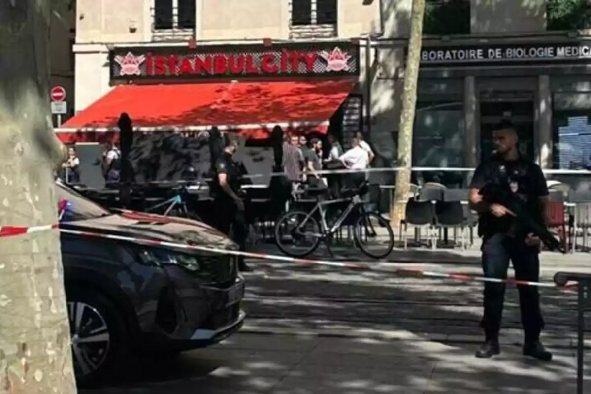 An armed police officer guards a cordoned-off area in front of a restaurant with several people gathered on the terrace. An armed police officer guards a cordoned-off area in front of a restaurant with several people gathered on the terrace.