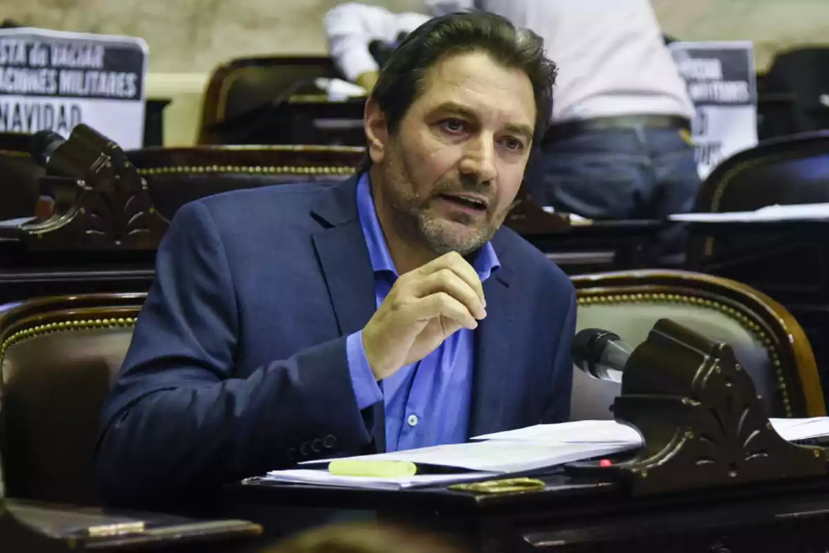 Brown-haired, bearded man speaking in a parliamentary chamber with papers and a microphone in front of him Brown-haired, bearded man speaking in a parliamentary chamber with papers and a microphone in front of him