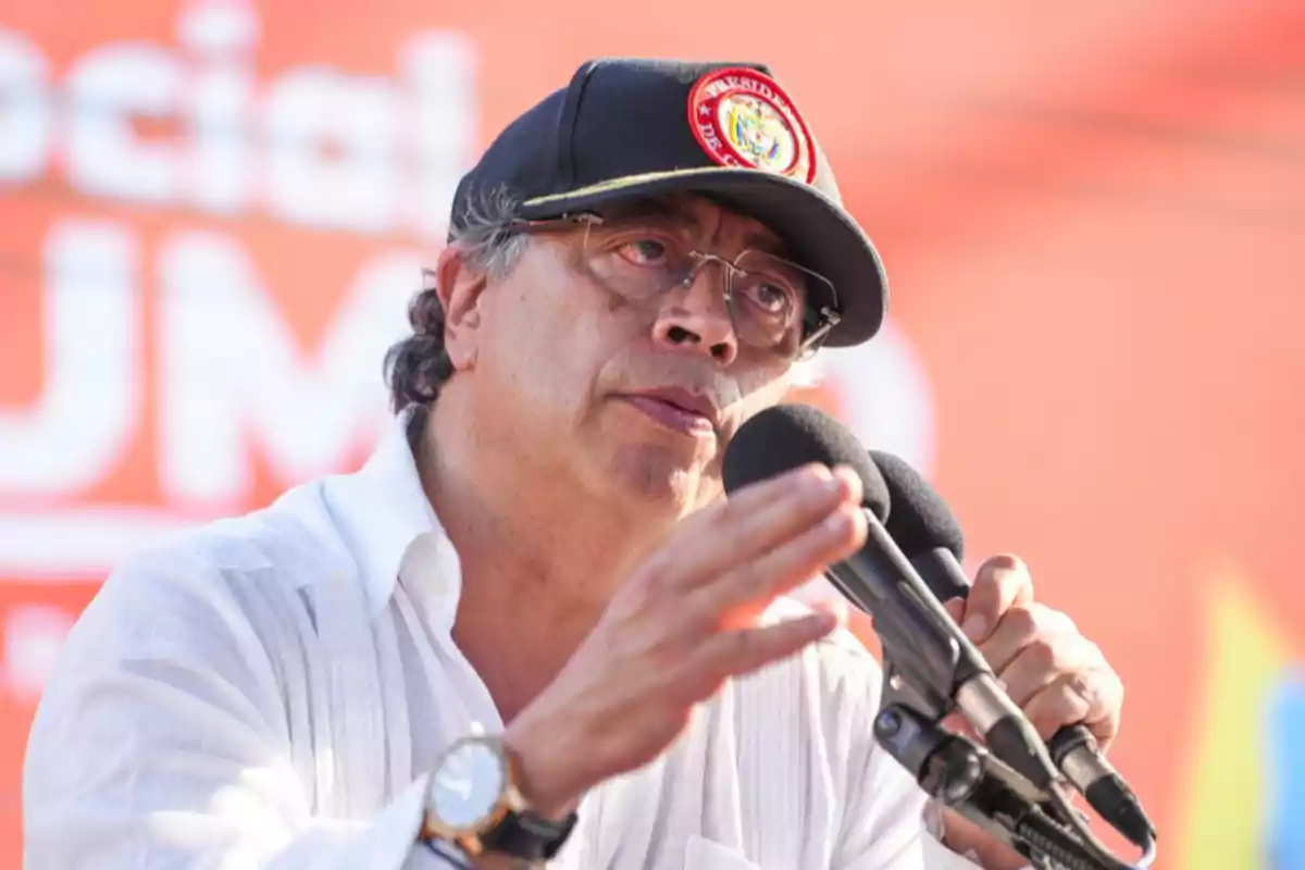Man with glasses and a dark cap speaking in front of two microphones while gesturing with his hand