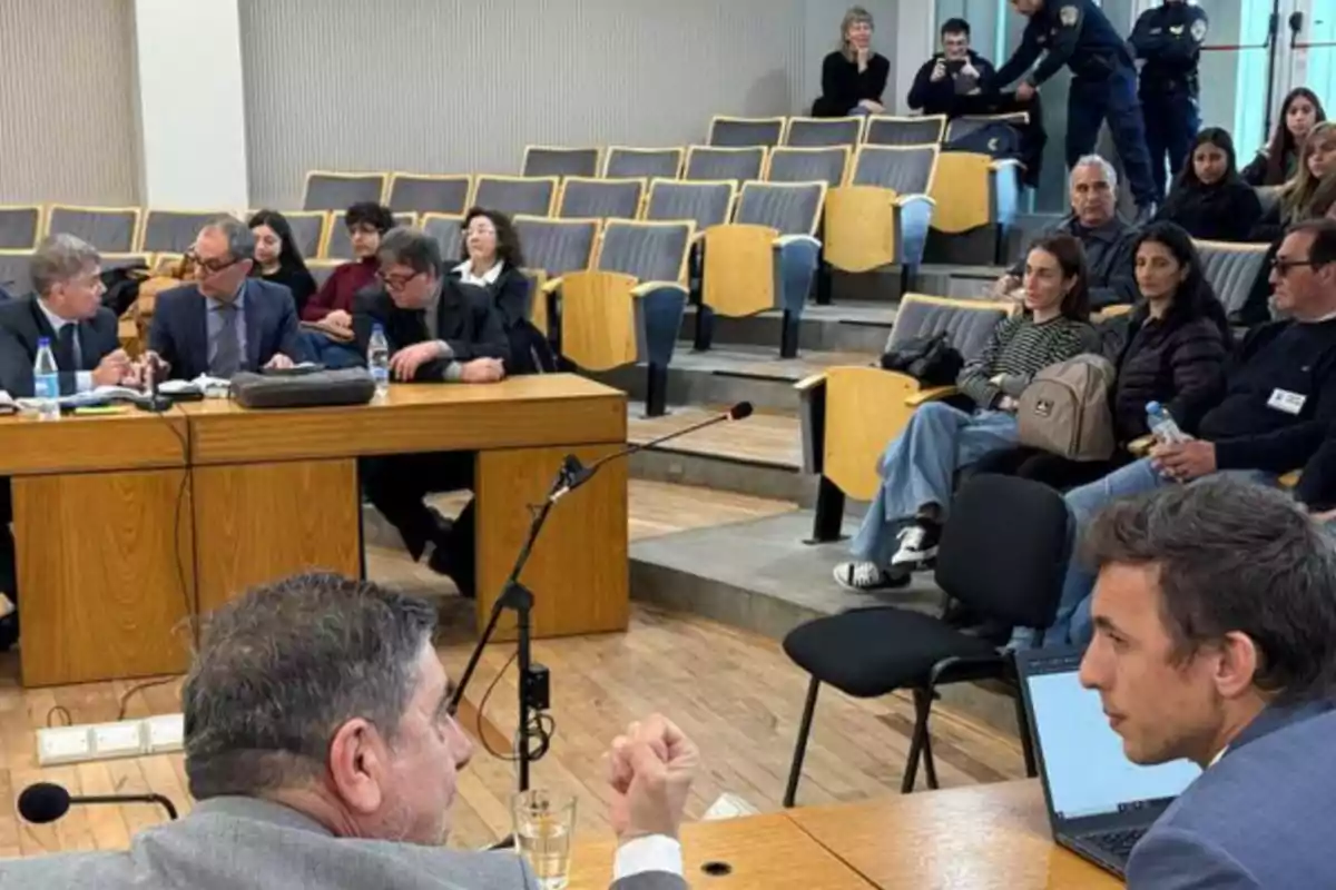 Personas sentadas en una sala de audiencias con bancas de madera y varias personas conversando en una mesa al frente Personas sentadas en una sala de audiencias con bancas de madera y varias personas conversando en una mesa al frente
