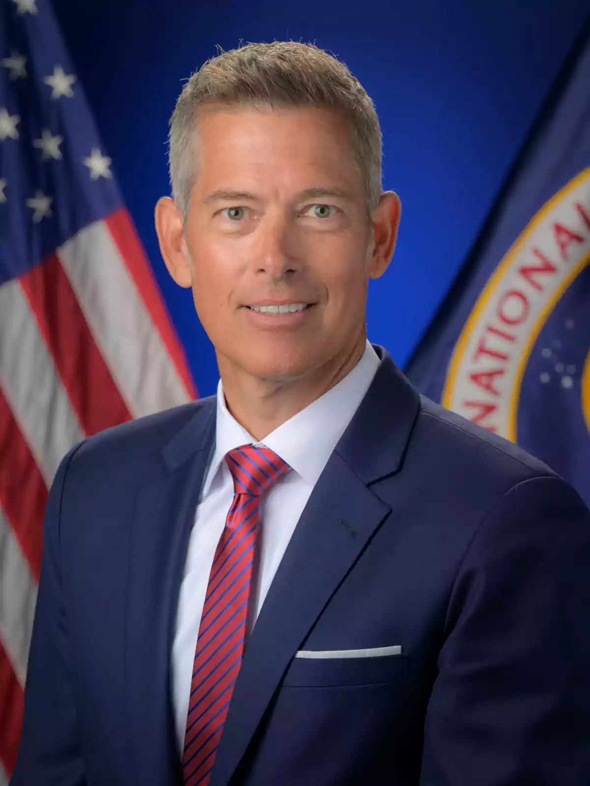 Short-haired, gray-haired man wearing a blue suit and red tie with a background of the American flag and an institutional emblem Short-haired, gray-haired man wearing a blue suit and red tie with a background of the American flag and an institutional emblem