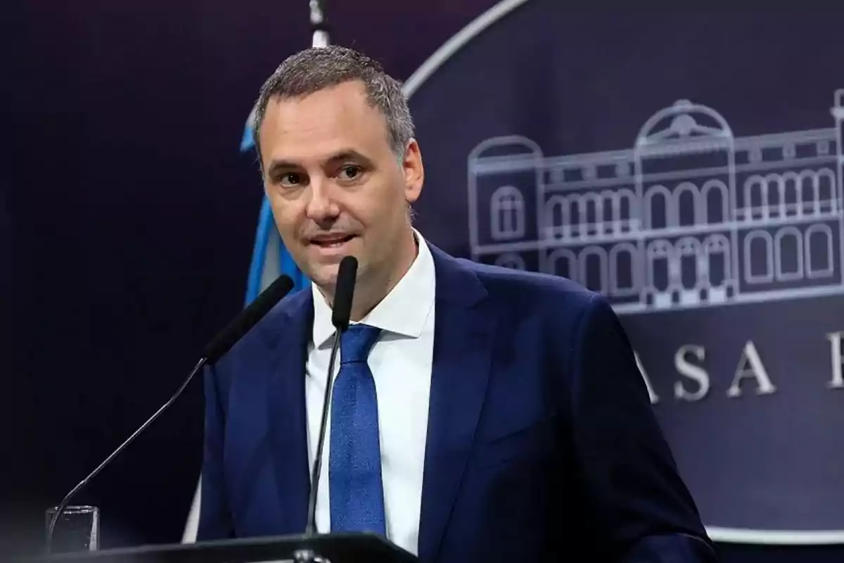 Man in a blue suit speaking in front of microphones at a press conference with an institutional background