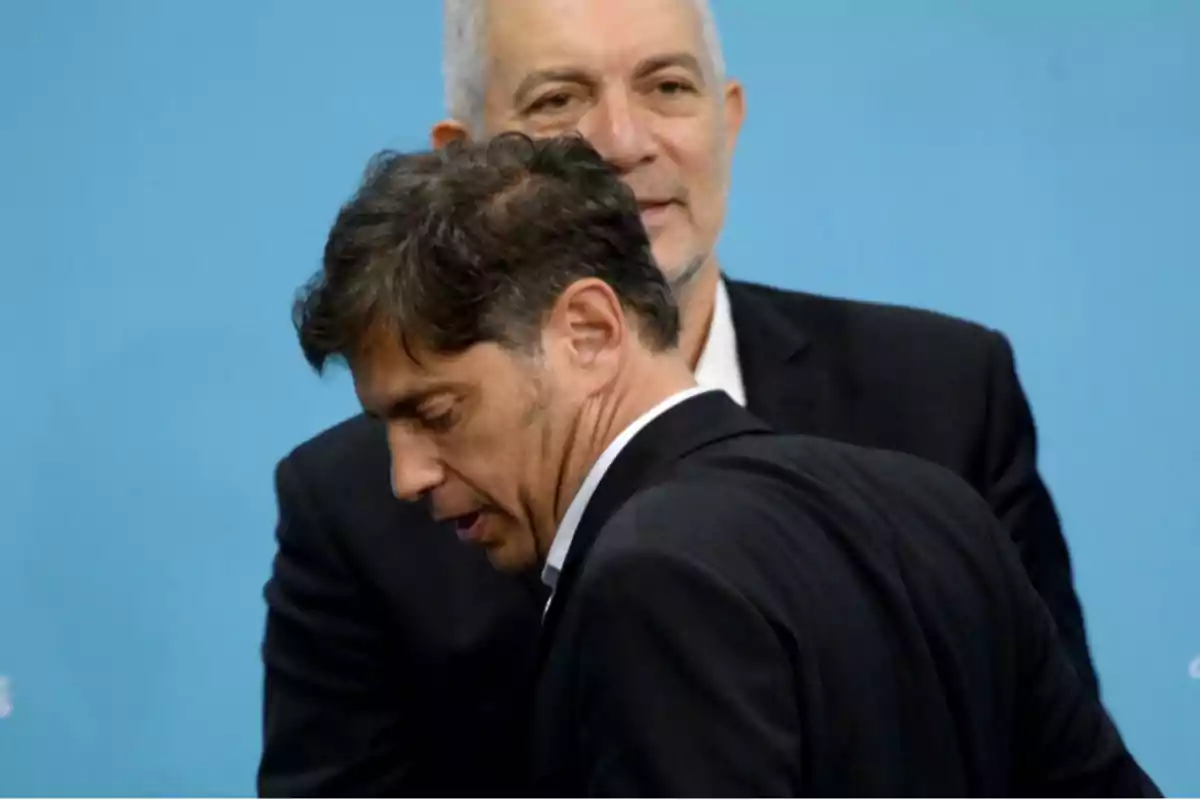 Two men in dark suits at a formal event, one of them in the foreground looking down while the other stands behind with a neutral expression against a blue background.