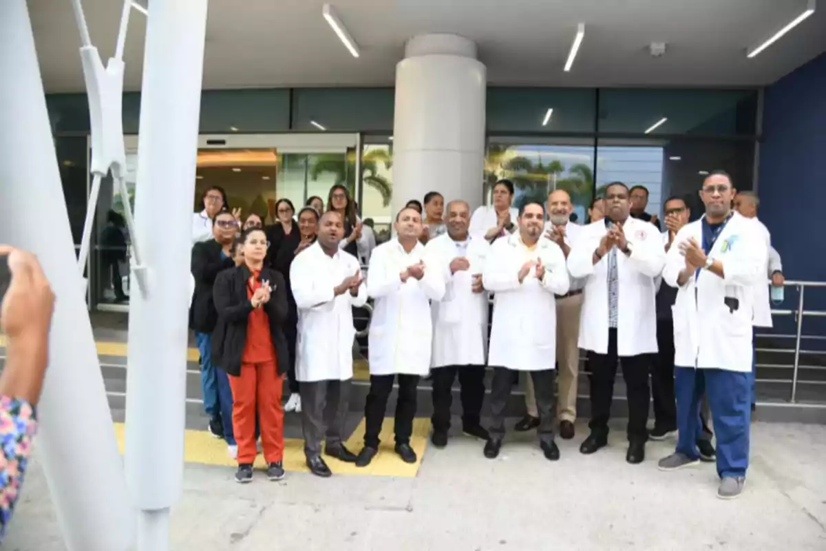 Grupo de médicos y personal de salud de pie frente a la entrada de un hospital, aplaudiendo y mirando hacia la cámara. Grupo de médicos y personal de salud de pie frente a la entrada de un hospital, aplaudiendo y mirando hacia la cámara.