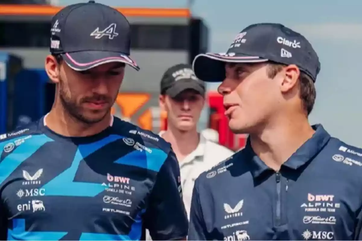 Two Formula 1 team drivers in blue uniforms and caps talk while walking in the paddock. Two Formula 1 team drivers in blue uniforms and caps talk while walking in the paddock.