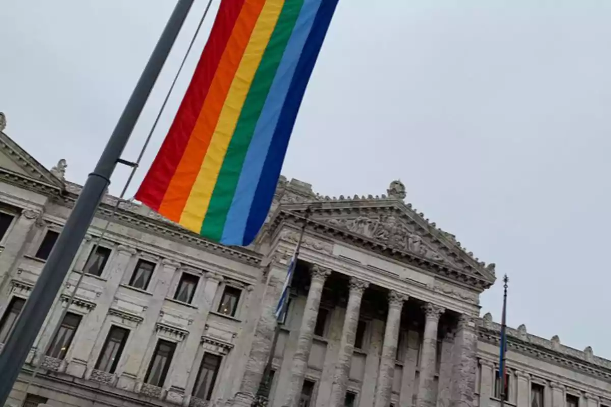 Bandera del orgullo LGBT ondeando frente a un edificio antiguo de arquitectura clásica con columnas y esculturas en la fachada
