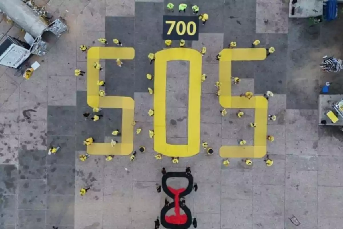 Aerial view of people forming the message SOS with large yellow letters and an hourglass on the ground, next to the number 700 at the top.