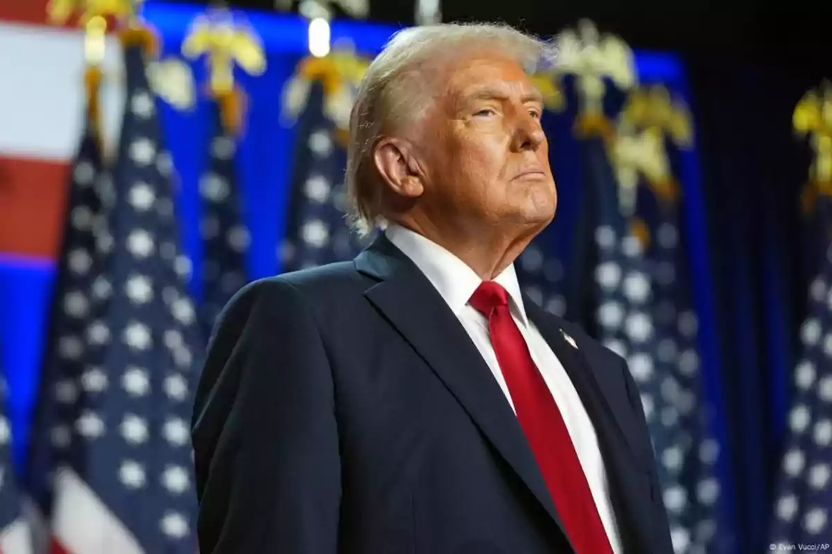 Blond-haired man in a dark suit and red tie posing in front of several United States flags