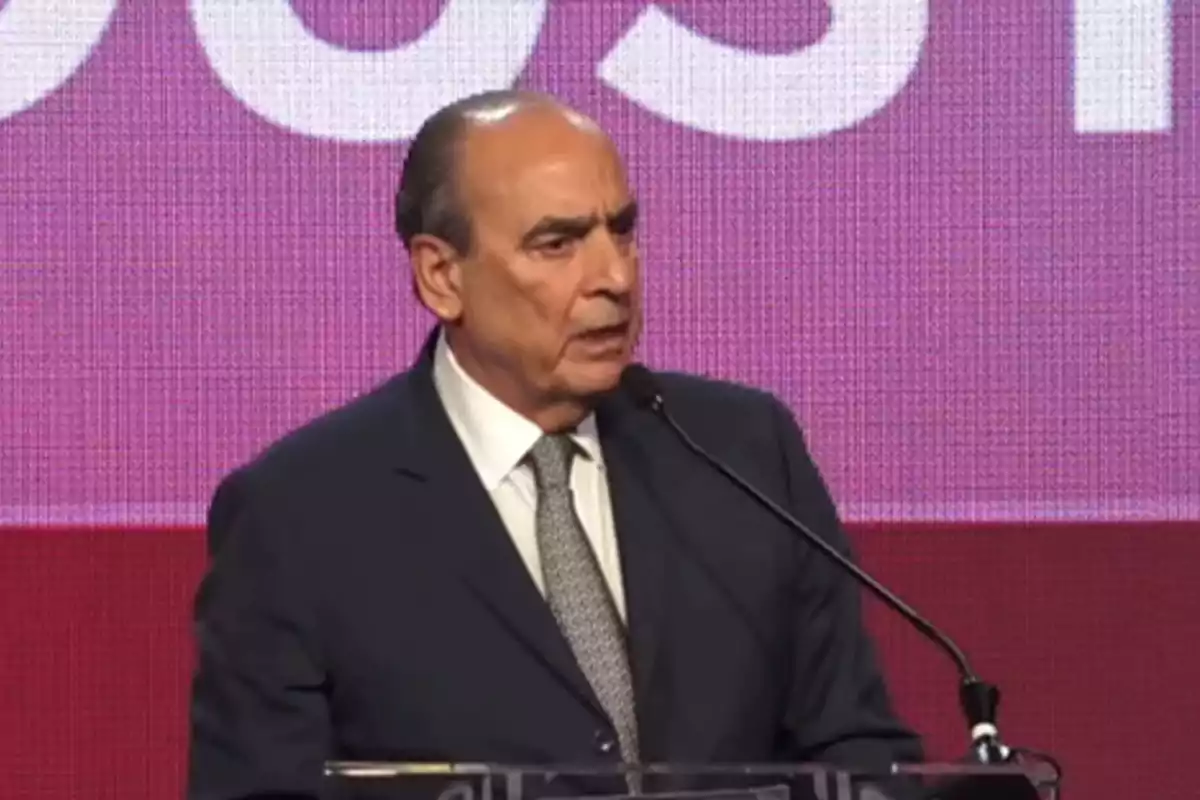 Man in a dark suit speaking at a podium with a microphone in front of a pink background screen