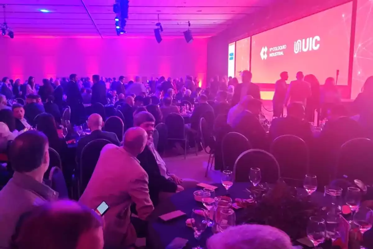 People sitting and standing in a room lit with purple and pink lights during a corporate event, with large screens in the background displaying logos and text. People sitting and standing in a room lit with purple and pink lights during a corporate event, with large screens in the background displaying logos and text.