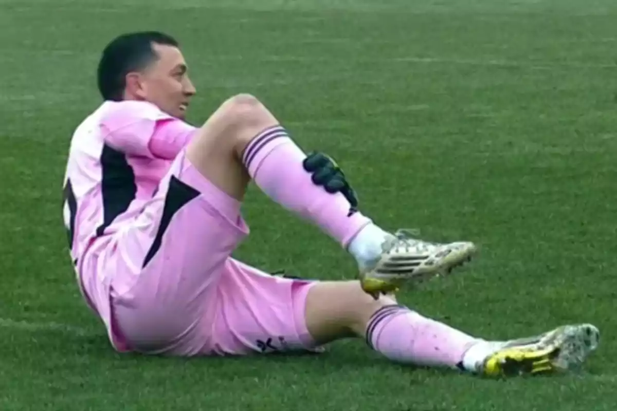 Goalkeeper in a pink uniform sitting on the grass holding his leg with one hand while appearing to be injured Goalkeeper in a pink uniform sitting on the grass holding his leg with one hand while appearing to be injured