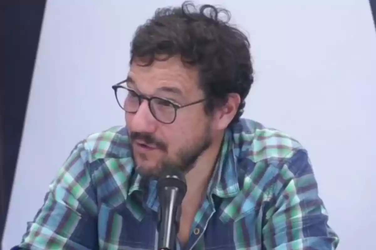 Man with glasses and a green plaid jersey speaking in front of a microphone Man with glasses and a green plaid jersey speaking in front of a microphone