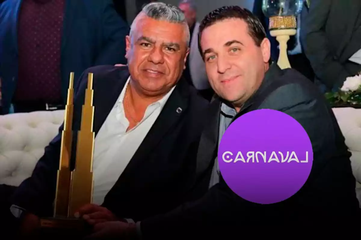 Dos hombres vestidos de traje oscuro posan sentados en un sofá blanco durante un evento, uno de ellos sostiene un trofeo dorado y hay un círculo violeta con la palabra "CARNAVAL" cubriendo parcialmente la imagen.