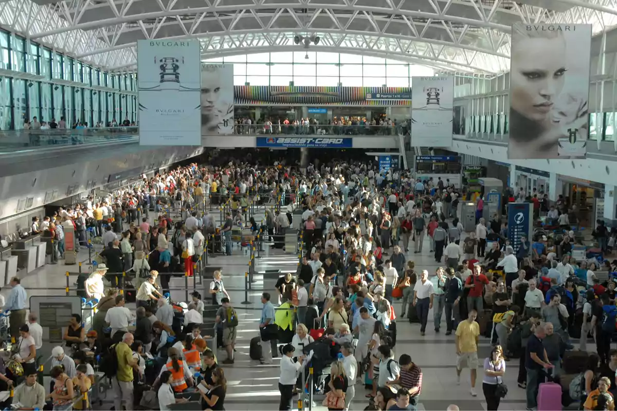 Airport terminal full of people standing in line and walking with suitcases under a metal-structured roof and large advertising signs Airport terminal full of people standing in line and walking with suitcases under a metal-structured roof and large advertising signs