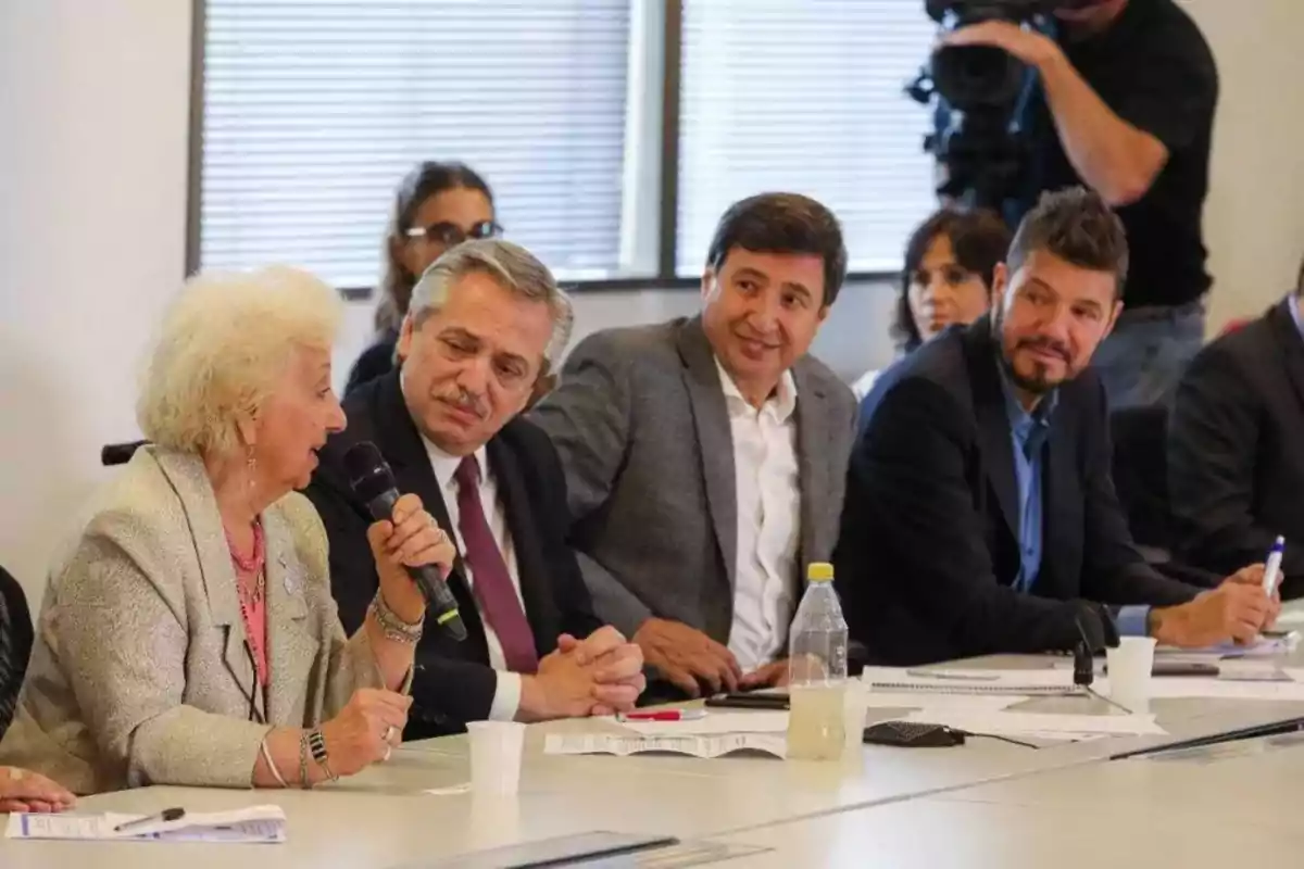 Varias personas sentadas alrededor de una mesa en una reunión, una mujer mayor habla por micrófono mientras los demás la escuchan atentamente Varias personas sentadas alrededor de una mesa en una reunión, una mujer mayor habla por micrófono mientras los demás la escuchan atentamente