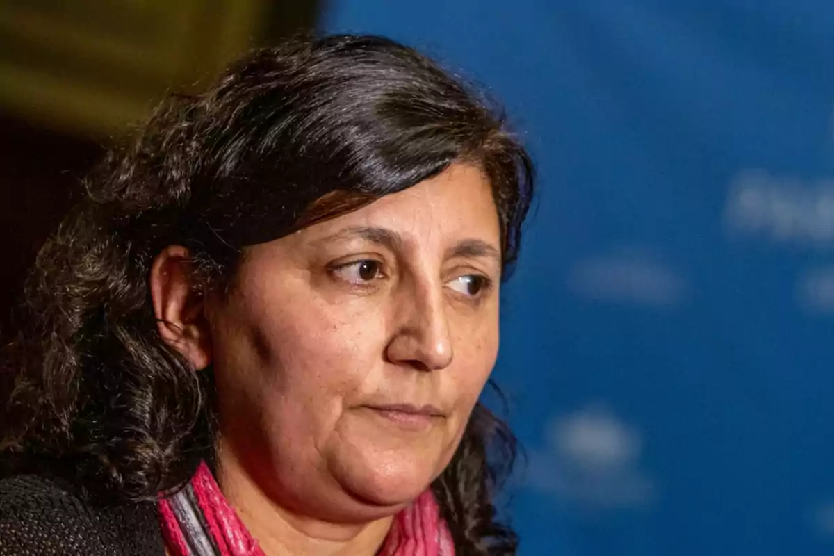 Dark-haired, wavy-haired woman with a pink scarf looking to the side against a blurred blue background