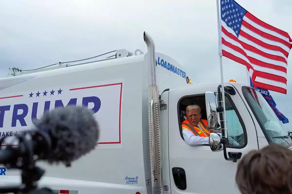 A man in an orange vest drives a white truck decorated with an American flag and a campaign sign, while being recorded by a camera.