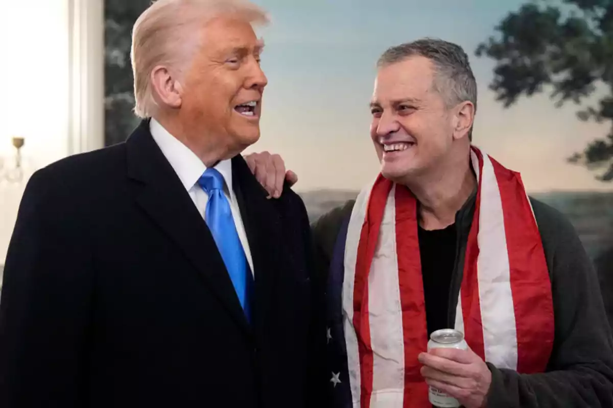 Dos hombres sonríen y conversan, uno lleva un traje con corbata azul y el otro tiene una bandera de Estados Unidos sobre los hombros y sostiene una lata Dos hombres sonríen y conversan, uno lleva un traje con corbata azul y el otro tiene una bandera de Estados Unidos sobre los hombros y sostiene una lata