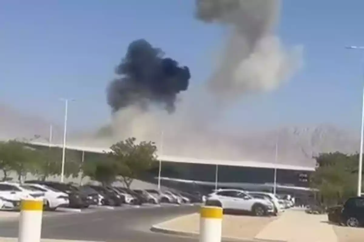 Humo negro y blanco elevándose desde un edificio detrás de un estacionamiento lleno de autos en un día soleado