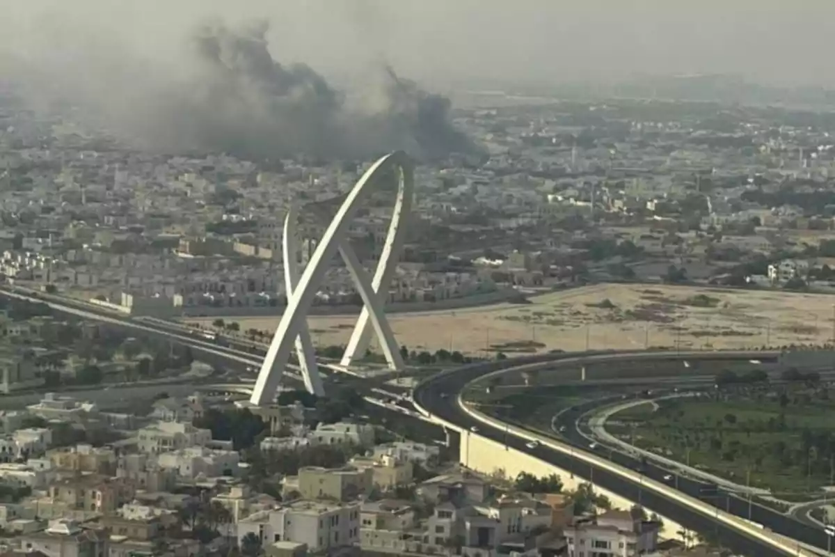 Vista panorámica de una ciudad con un monumento moderno en forma de arco doble y una columna de humo negro al fondo