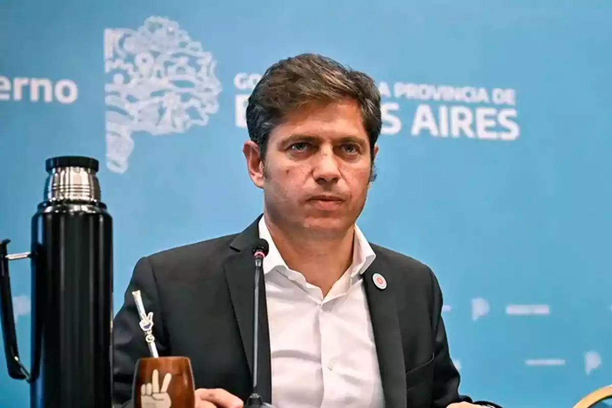 Hombre de cabello corto y saco oscuro sentado frente a un micrófono con un termo y un mate sobre la mesa y un fondo azul con texto borroso
