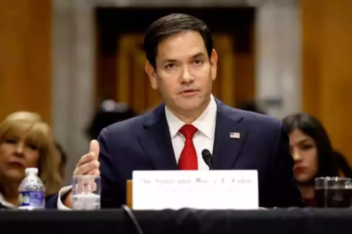 Dark-haired man in a blue suit and red tie speaking into a microphone during a hearing or conference, with people seated in the background Dark-haired man in a blue suit and red tie speaking into a microphone during a hearing or conference, with people seated in the background