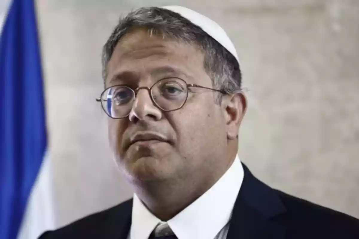 Man with glasses and a white kippah wearing a dark suit and tie in front of a light-colored wall