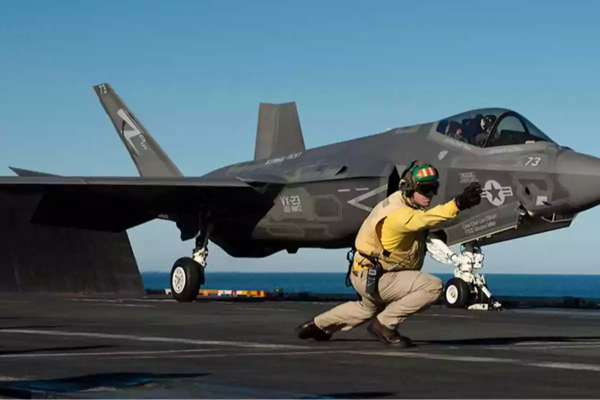 Un avión de combate en la cubierta de un portaaviones con un tripulante señalando hacia adelante Un avión de combate en la cubierta de un portaaviones con un tripulante señalando hacia adelante
