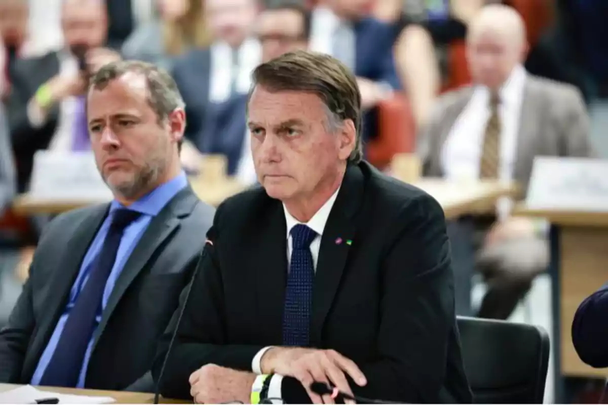 Two men in suits sitting at a table during a formal meeting with blurred people in the background