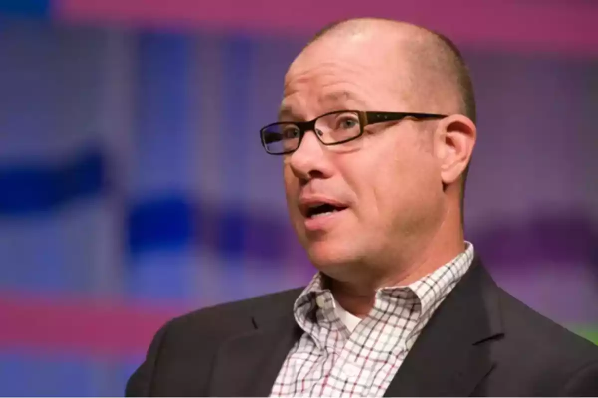 Bald man with glasses and a dark jacket speaking, with a blurred blue and pink background Bald man with glasses and a dark jacket speaking, with a blurred blue and pink background