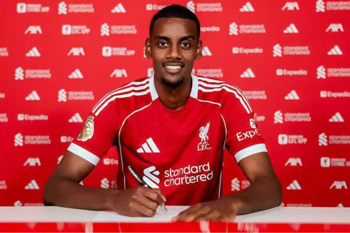 Soccer player wearing a Liverpool jersey signing a contract in front of a red background with club and sponsor logos