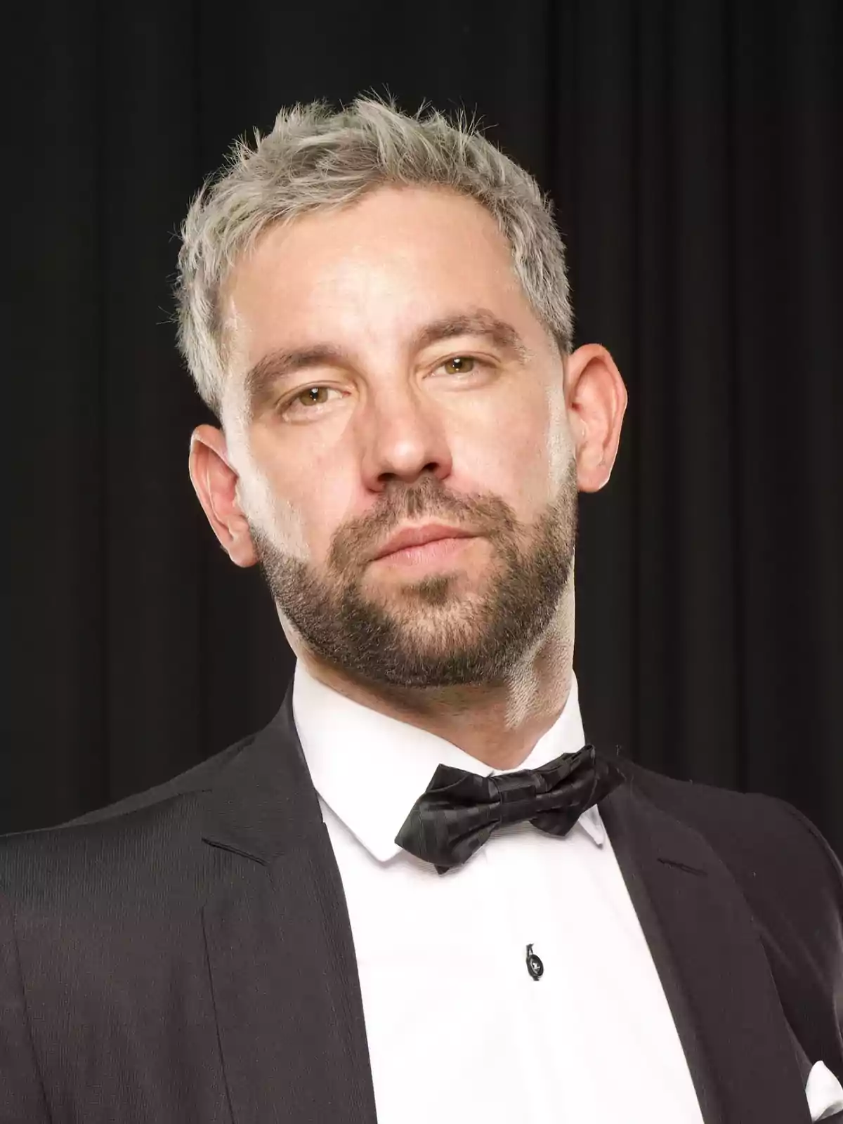 Hombre de cabello corto y barba vestido con esmoquin negro y pajarita frente a un fondo oscuro Hombre de cabello corto y barba vestido con esmoquin negro y pajarita frente a un fondo oscuro