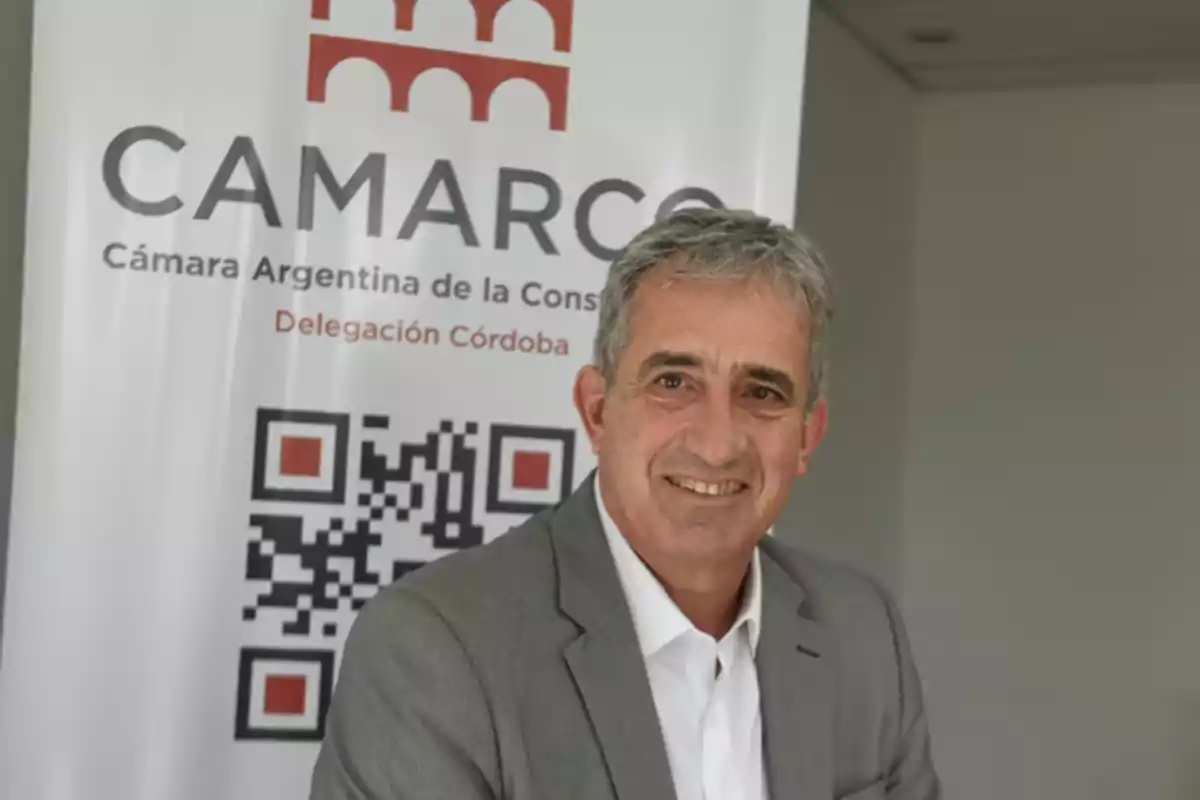 Hombre de cabello canoso con saco gris y camisa blanca sonriendo frente a un cartel de la Cámara Argentina de la Construcción Delegación Córdoba Hombre de cabello canoso con saco gris y camisa blanca sonriendo frente a un cartel de la Cámara Argentina de la Construcción Delegación Córdoba