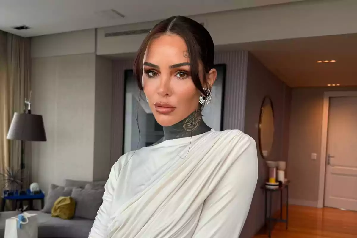 Mujer de cabello oscuro recogido con vestido blanco elegante y tatuaje en el cuello posando en una sala moderna