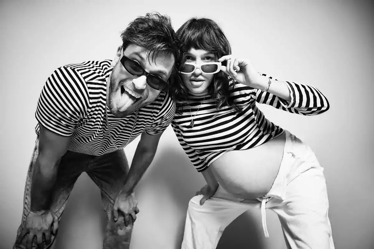 Pareja divertida con camisetas de rayas y gafas de sol posando juntos, la mujer muestra su embarazo y ambos sonríen en actitud juguetona Pareja divertida con camisetas de rayas y gafas de sol posando juntos, la mujer muestra su embarazo y ambos sonríen en actitud juguetona