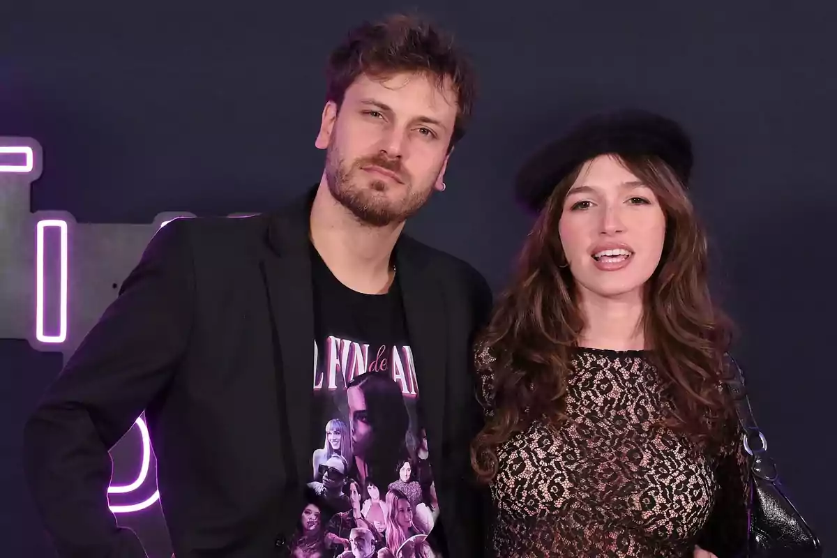 Pareja posando en un evento con fondo oscuro y luces de neón, él lleva saco negro y camiseta estampada, ella viste blusa de encaje negra y boina