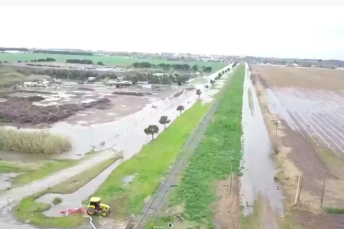 Aerial view of a flooded rural area with waterlogged fields, a road and railroad tracks partially covered by water, and a yellow tractor in the foreground. Aerial view of a flooded rural area with waterlogged fields, a road and railroad tracks partially covered by water, and a yellow tractor in the foreground.