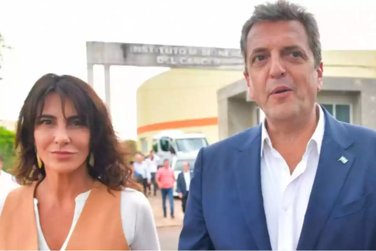 A woman and a man dressed formally are outdoors in front of a building with a blurry sign and several people in the background. A woman and a man dressed formally are outdoors in front of a building with a blurry sign and several people in the background.