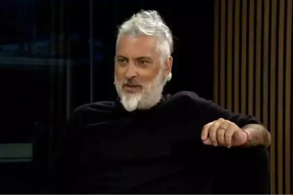 Hombre de cabello y barba canosos sentado en un sillón negro con fondo oscuro y líneas verticales de madera Hombre de cabello y barba canosos sentado en un sillón negro con fondo oscuro y líneas verticales de madera