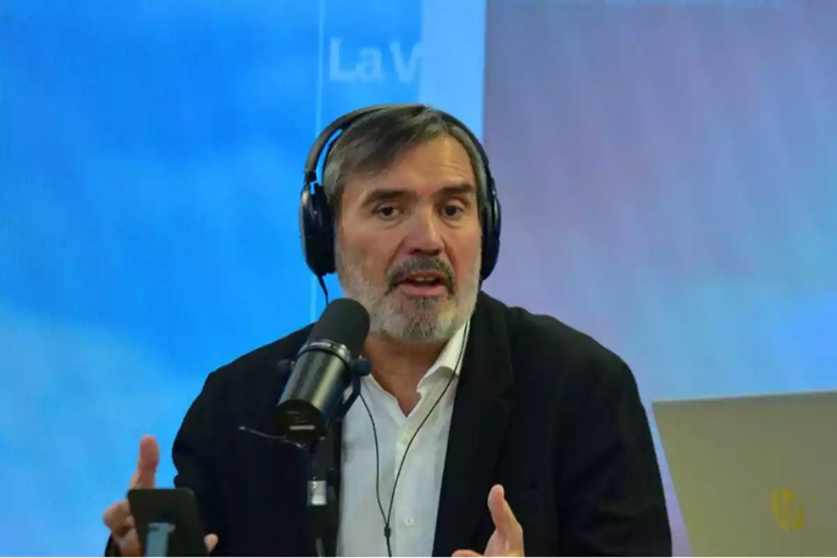 Hombre de mediana edad con audífonos hablando frente a un micrófono en un estudio de radio o podcast, con una laptop a un lado y fondo azul difuminado. Hombre de mediana edad con audífonos hablando frente a un micrófono en un estudio de radio o podcast, con una laptop a un lado y fondo azul difuminado.