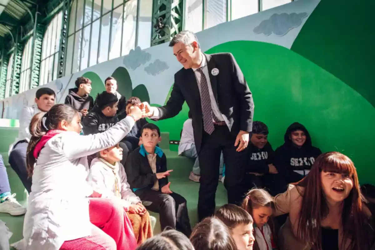A man in a suit greets a girl with a fist bump while several children and adults watch and smile in a colorful, bright space.