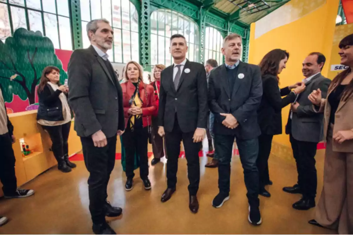 A group of people dressed in formal and casual attire is gathered in a colorful indoor space with yellow and green walls; some are talking while others are looking at the camera. A group of people dressed in formal and casual attire is gathered in a colorful indoor space with yellow and green walls; some are talking while others are looking at the camera.