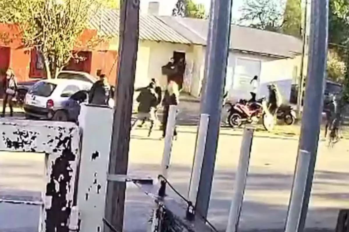 Several people run and gather on a street in front of a house while parked cars and motorcycles can be seen. Several people run and gather on a street in front of a house while parked cars and motorcycles can be seen.