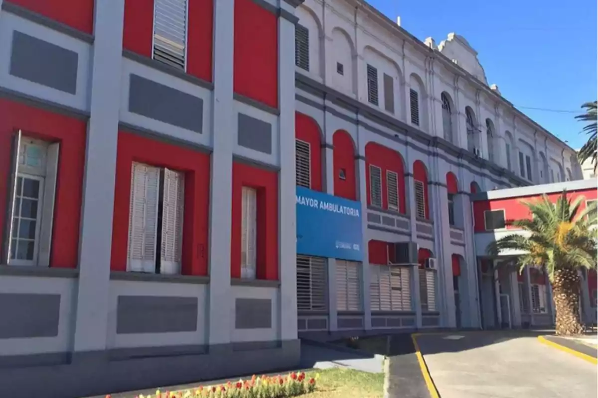 Fachada de un edificio hospitalario de dos pisos con paredes rojas y grises, ventanas con persianas y un cartel azul que indica área ambulatoria, junto a una palmera y un camino de acceso. Fachada de un edificio hospitalario de dos pisos con paredes rojas y grises, ventanas con persianas y un cartel azul que indica área ambulatoria, junto a una palmera y un camino de acceso.