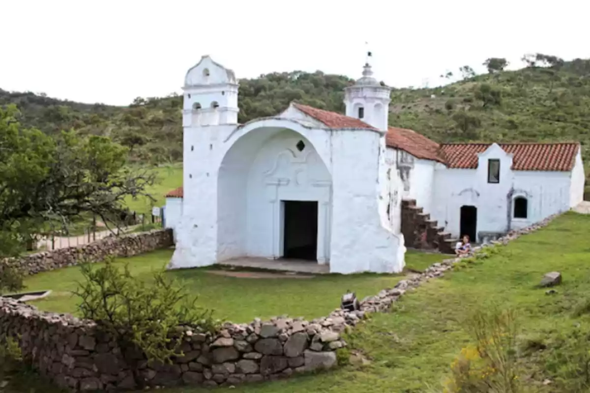 Iglesia colonial blanca con techo de tejas rojas rodeada de un muro de piedra y vegetación en un entorno rural Iglesia colonial blanca con techo de tejas rojas rodeada de un muro de piedra y vegetación en un entorno rural